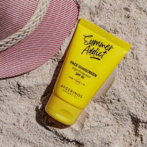 Жълта тубичка слънцезащитен крем за лице SPF 50 Avgerinos, легнала върху фин морски пясък до раирана сламена шапка